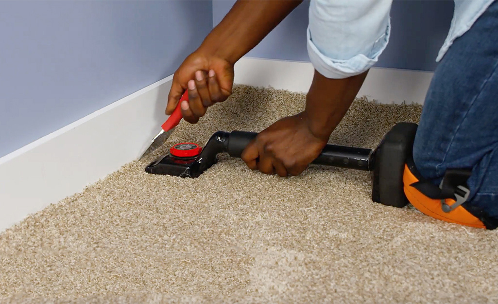 A person kneels on a knee kicker to anchor beige carpet along the baseboard of a room during installation.