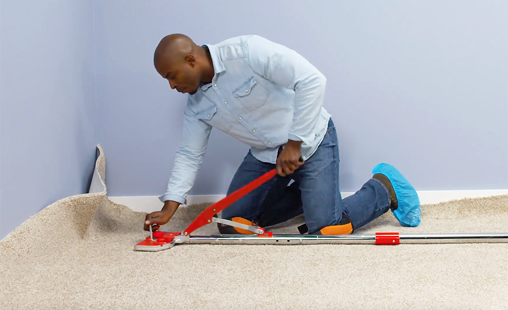 A man wearing a denim shirt, jeans and knee pads kneels on the floor to use a carpet stretcher as he installs beige carpet.