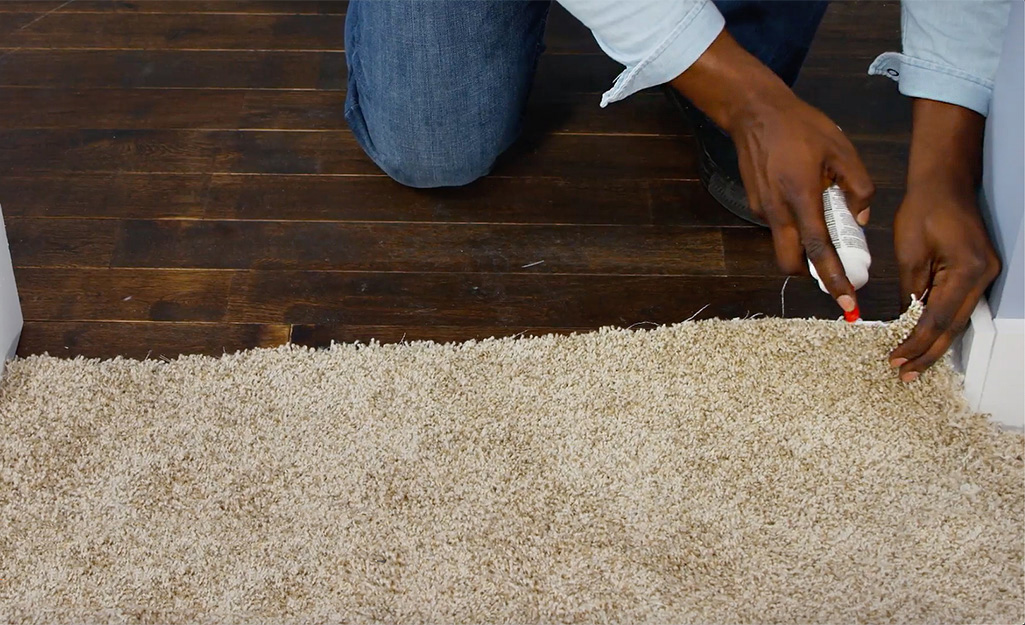 A person kneels to apply latex seam sealer underneath the rough edge of newly installed carpet where it meets a hardwood floor.