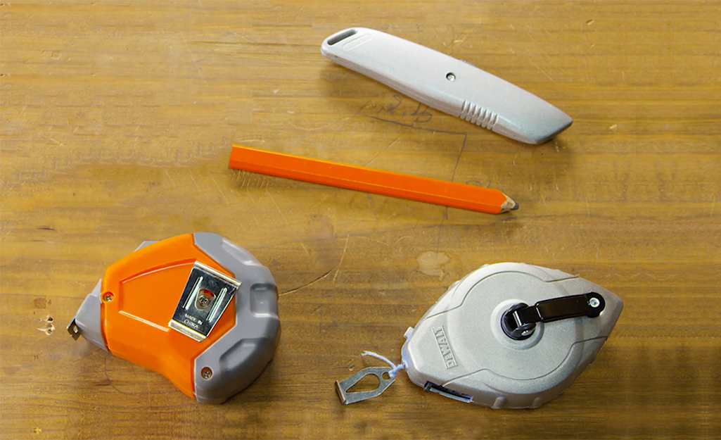 A measuring tape, pencil, chalk reel and utility knife lay on a plywood subfloor.