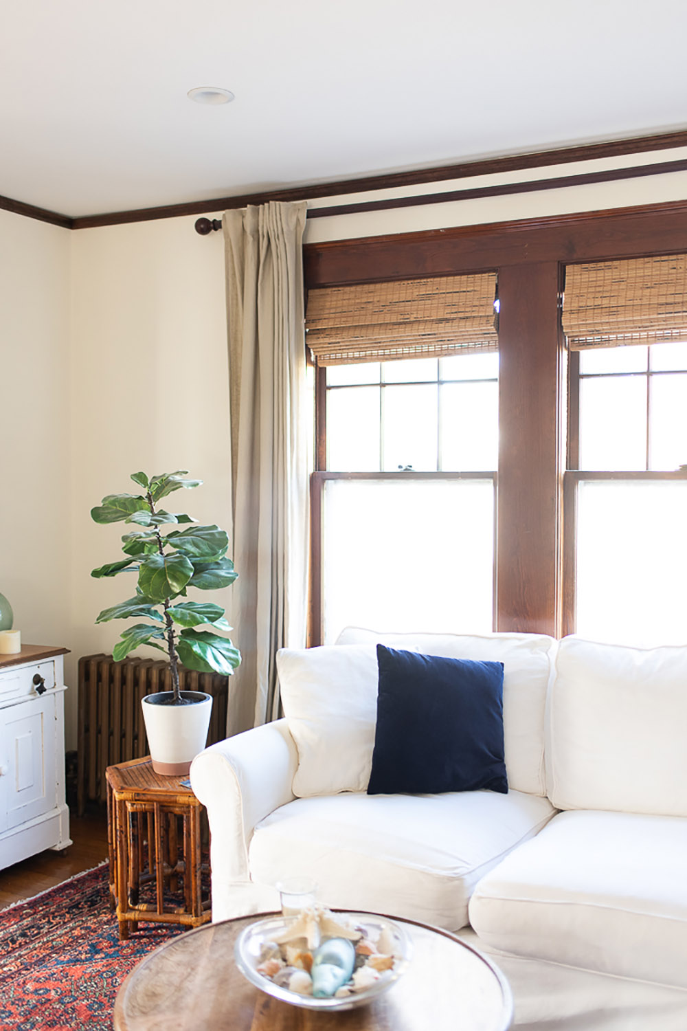 A white couch with a navy pillow sits in front of a window with woven roman shades.