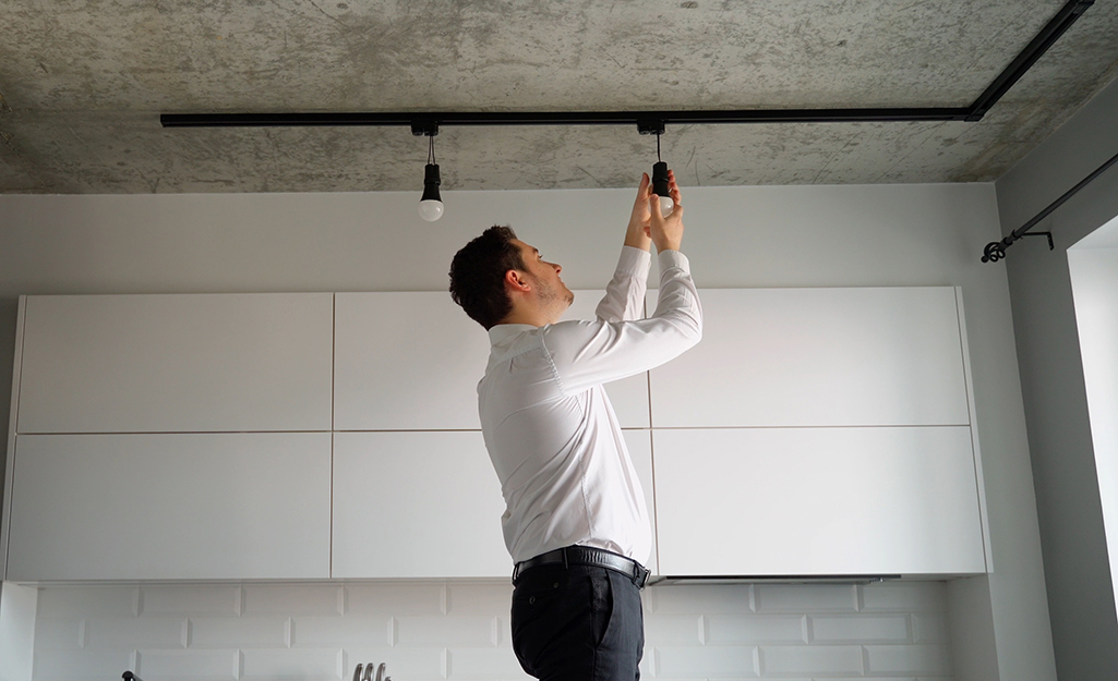 A man adjusts one of the fixtures of track lighting in front of a row of white cabinets.
