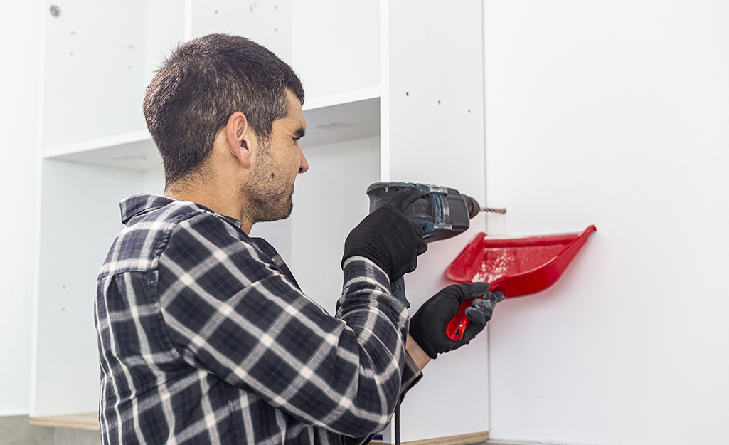 A person drilling a hole in a wall next to kitchen cabinet. A person drilling a hole in a wall next to kitchen cabinet.