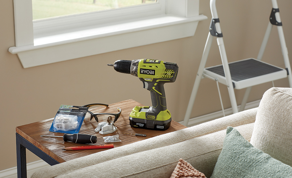 A drill, window lock, screwdriver, pencil and other tools needed to install a window lock sit on a table next to a stepladder.