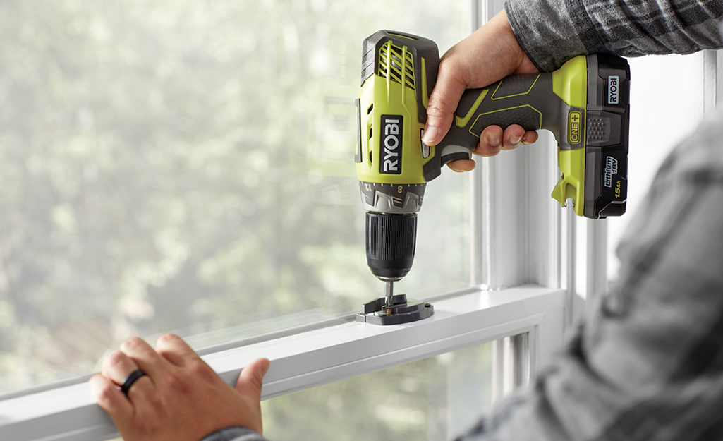 A person uses a drill to unscrew a window latch.