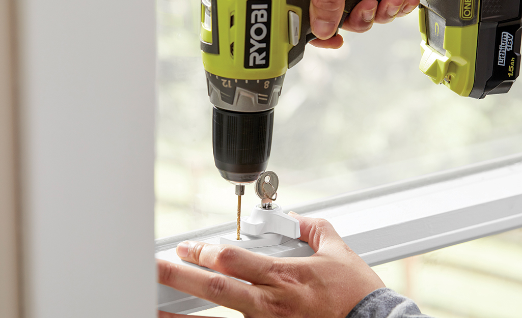 A person uses a drill while installing a window lock with a key.
