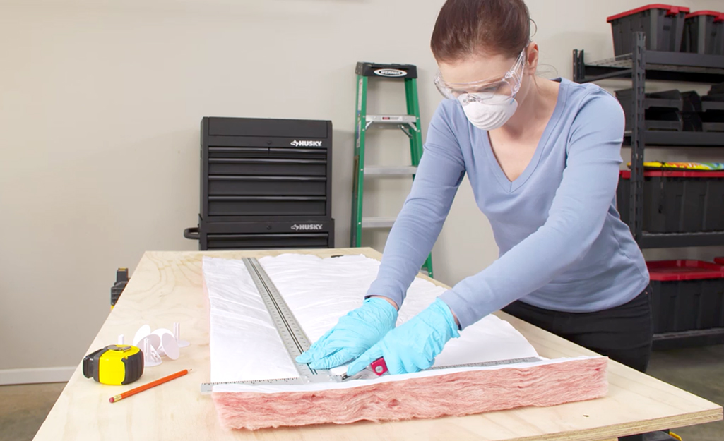 A person wearing protective gloves, glasses and a dust mask measuring and cutting fiberglass insulation.