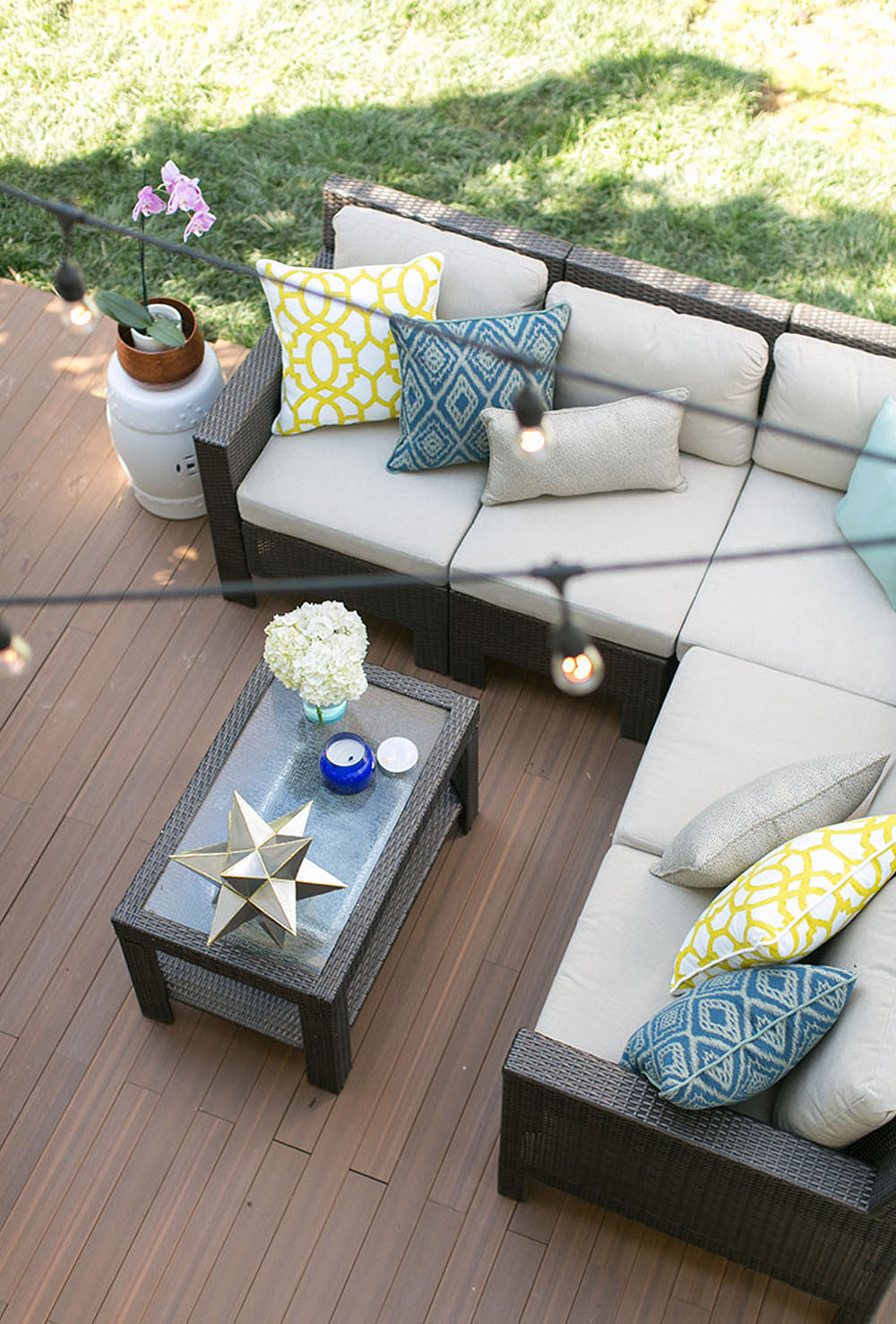 A deck floor decorated with an outdoor sectional, coffee table, pillows, and string lights.