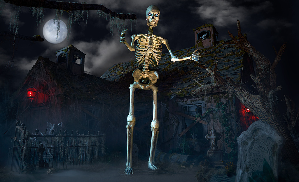 A 12-foot skeleton stands in front of a spooky house at night with a full moon in the sky.