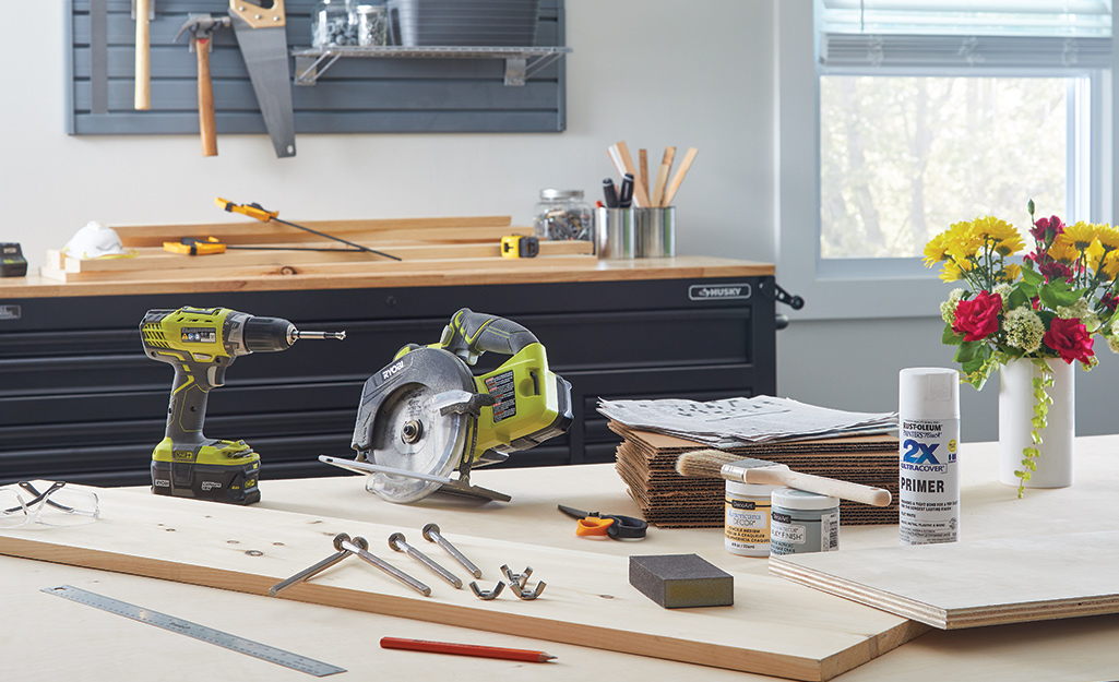 A drill, a circular saw, a stack of cardboard and newspaper and other supplies to make a flower press lay on a worktable.