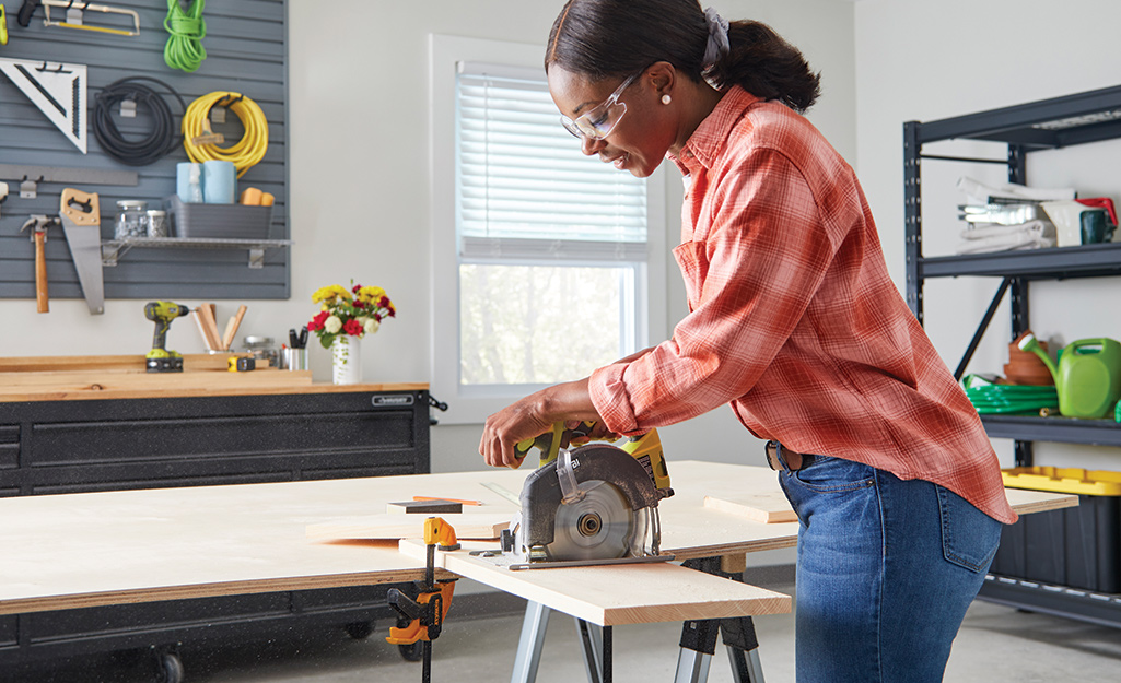 A woman wearing safety glasses cuts a piece of wood with a circular saw. 