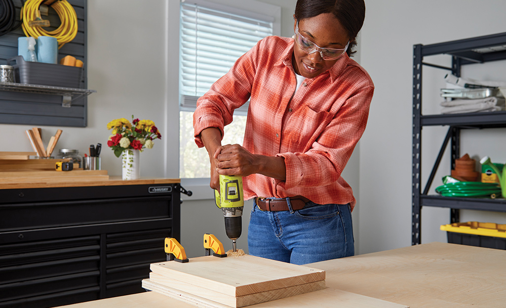 A woman wearing safety glasses drills a hole in the corner of two pieces of wood held together with clamps.