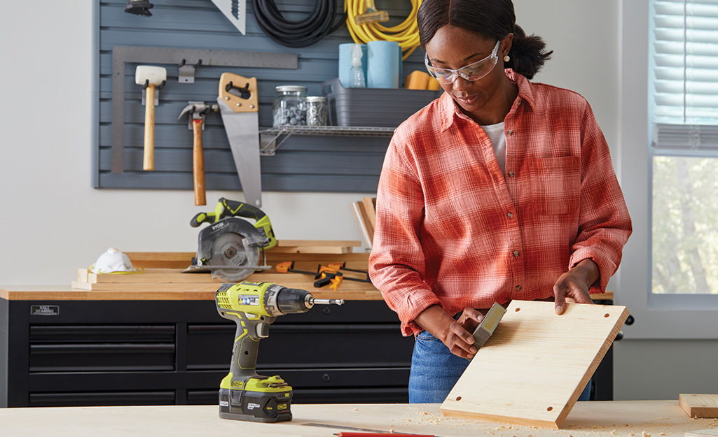 A woman in a workshop sands a square piece of wood that has a hole in each corner.