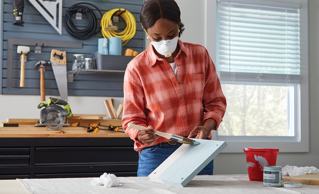 A woman wearing a mask applies paint to a square piece of wood with a brush.