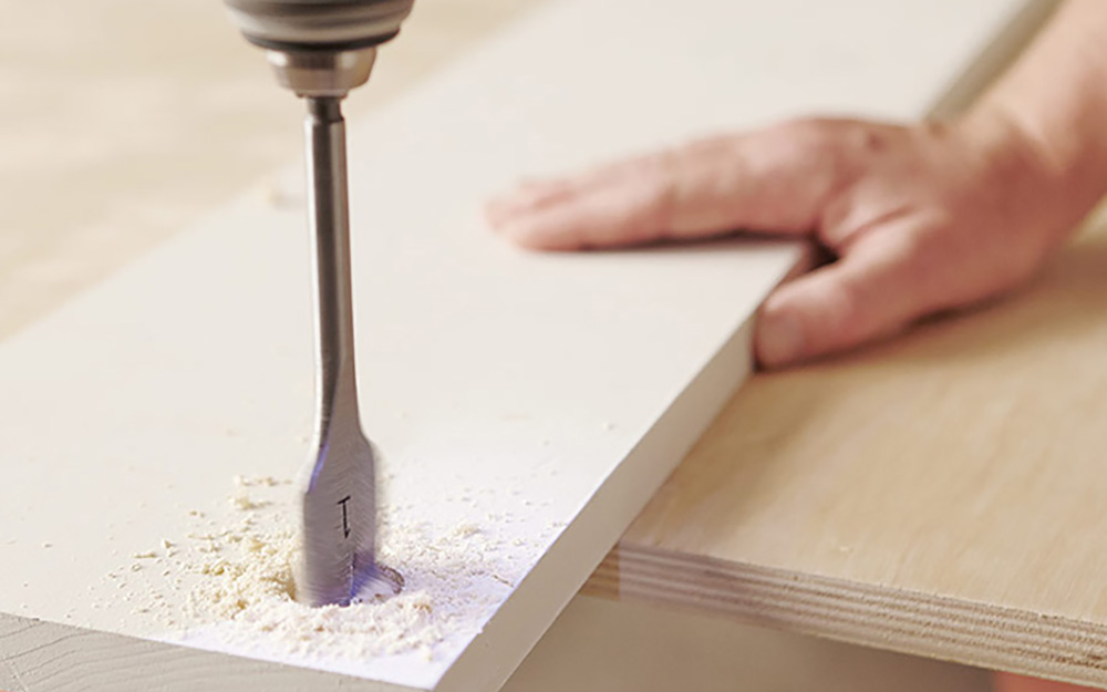 A person drills into a painted wood board with a spade bit.