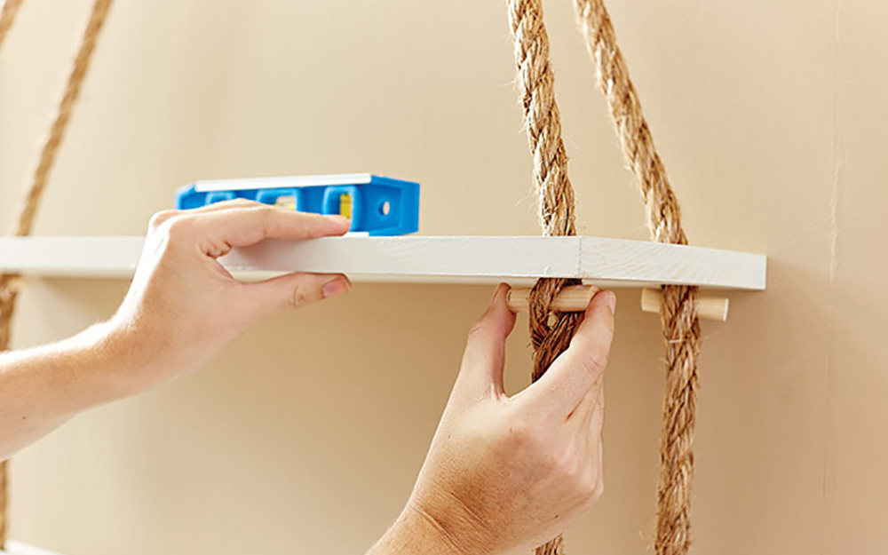 A person inserting dowels into the rope and checking the level of the shelf.