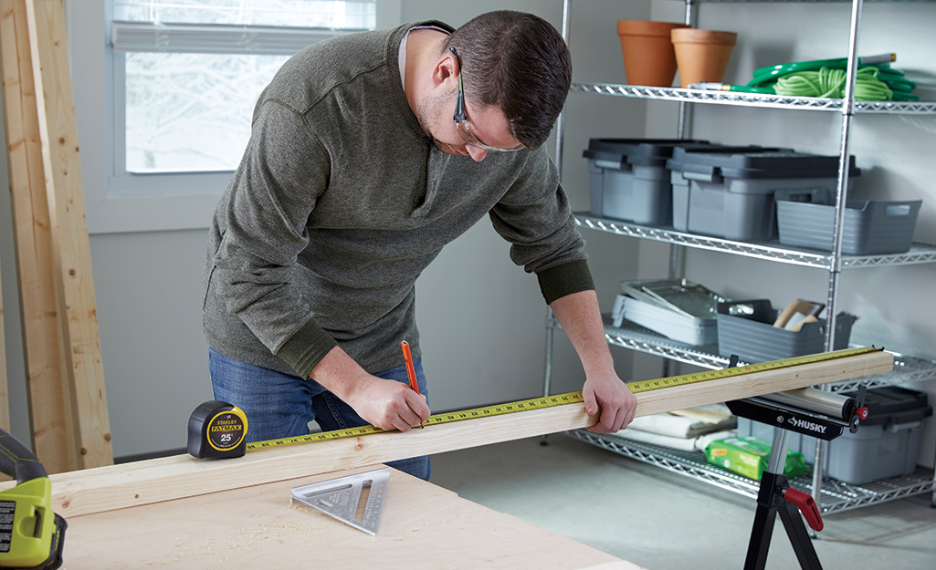 A man in a workshop uses a measuring tape and a pencil to mark where a board should be cut.