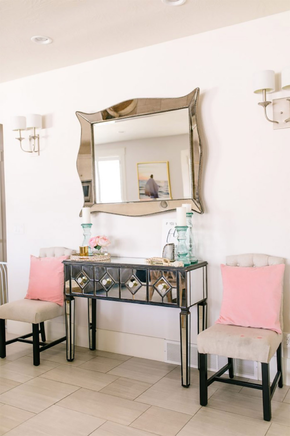 A black and mirrored console table with fabric chairs and pink pillows on either side.