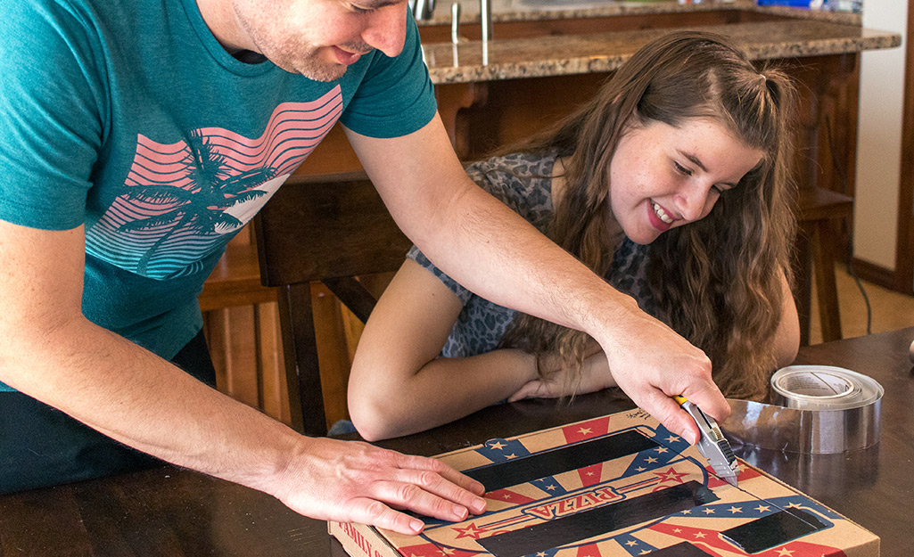 A man cutting into a pizza box while a young girl watches.