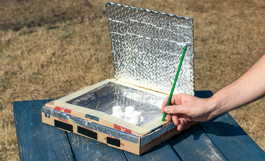A person placing a pencil to prop open a lid of a homemade solar oven.