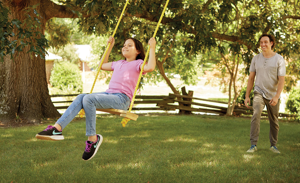 A girl wearing a pink shirt swings on a tree swing and a man stands behind the swing.