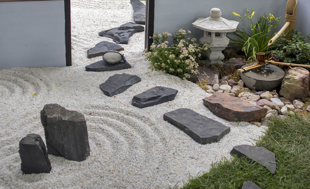 A Zen garden with circles traced in crushed white gravel, a stone lantern, some plants and black rocks and stepping stones.