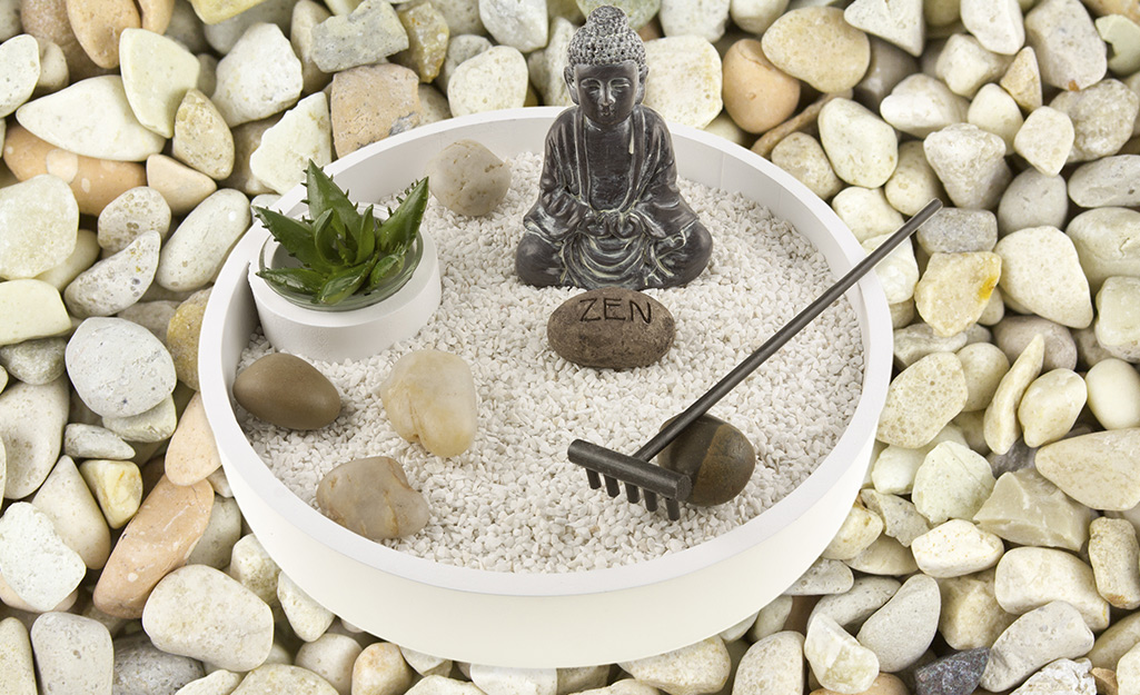 A mini Zen garden with sand, pebbles, a succulent, a small rake and a small Buddha figurine.