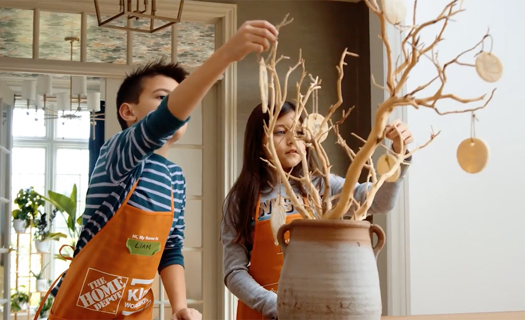 Kids wearing orange aprons from The Home Depot hang leaf ornaments on decorative tree branches in a vase.