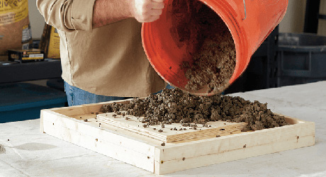 Someone pouring ready-mix concrete from a bucket into a plywood paver mold.