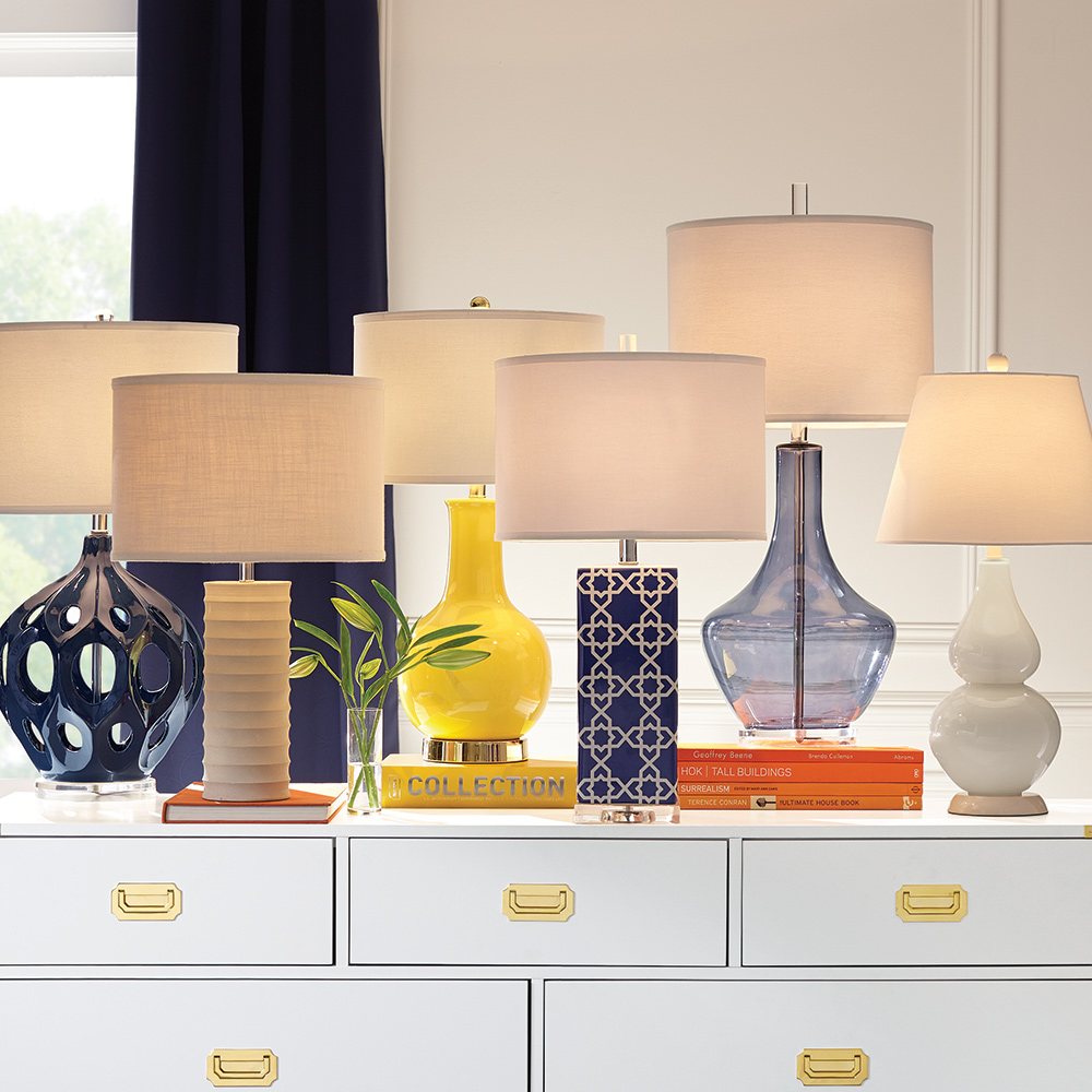An assortment of six table lamps sit side by side on a white dresser.