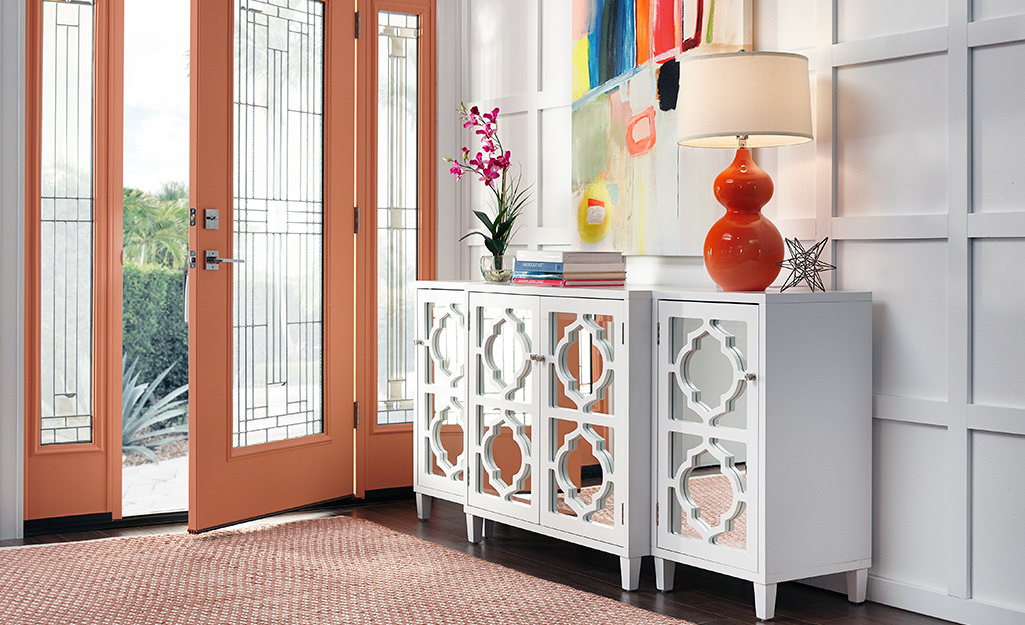 A home entryway features a white side table with mirrored doors and a bright red lamp with a simple barrel lamp shade.