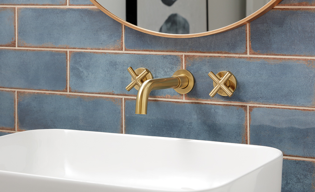 Gold wall-mounted bathroom sink fixtures are installed on a blue-tiled wall above a white vessel sink.