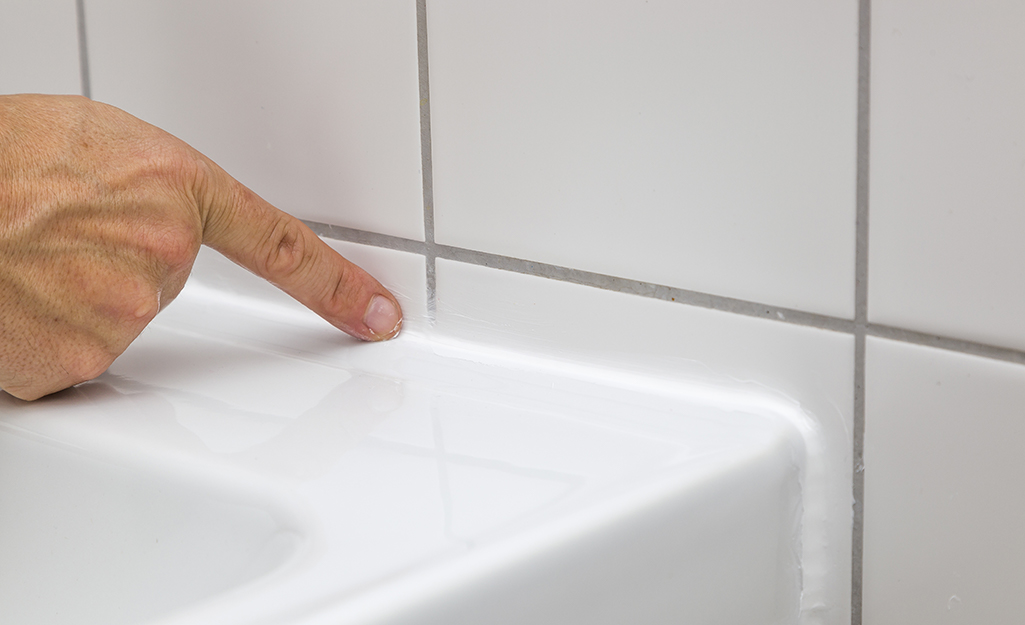 A finger points to caulk in the joint between a sink and tile wall.