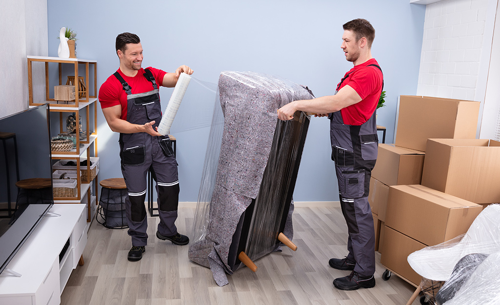Two professional movers wrap a couch.