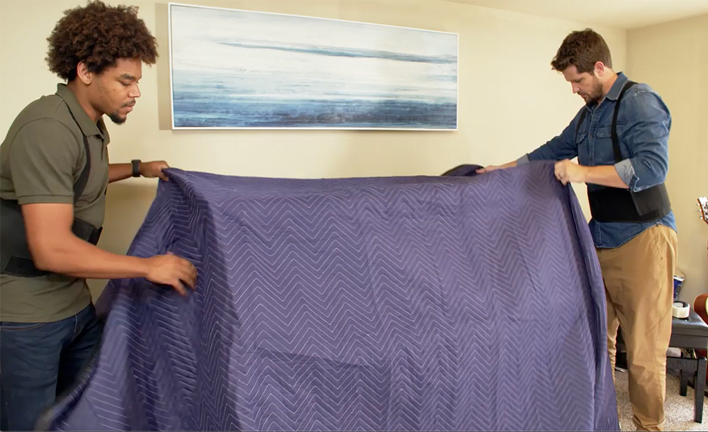Two men cover an upright piano with a moving blanket. 