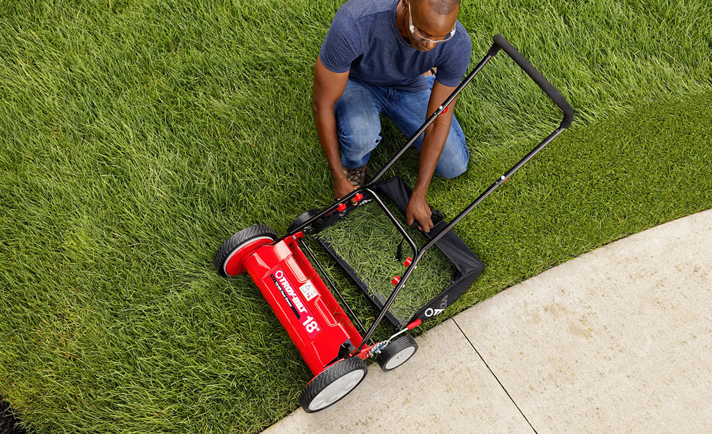 A man kneels on a lawn next to a red reel mower.