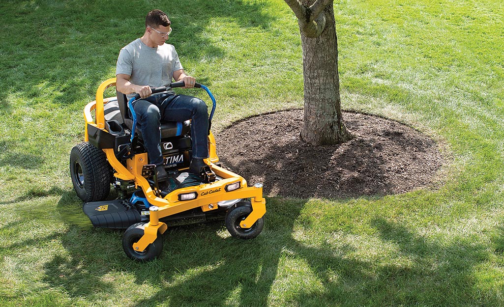 A man cuts the grass around a the base of a tree with a zero turn mower.