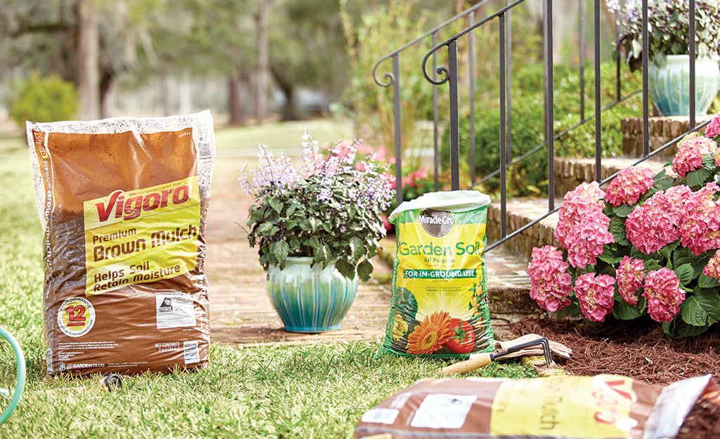 Bags of mulch and soil by pink flowers planted in a mulched garden bed.