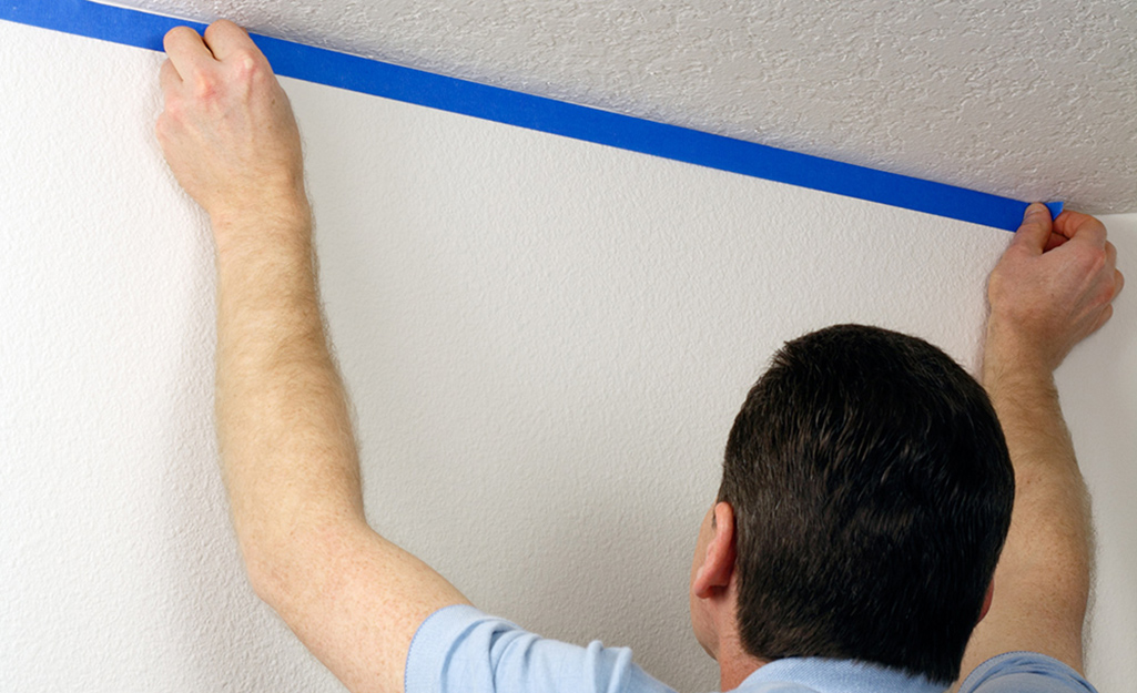 A man uses painter's tape to prep the walls before painting the ceiling.