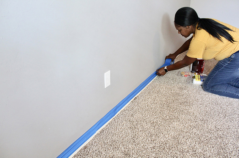 A woman taping the edges of a baseboard with painter's tape.