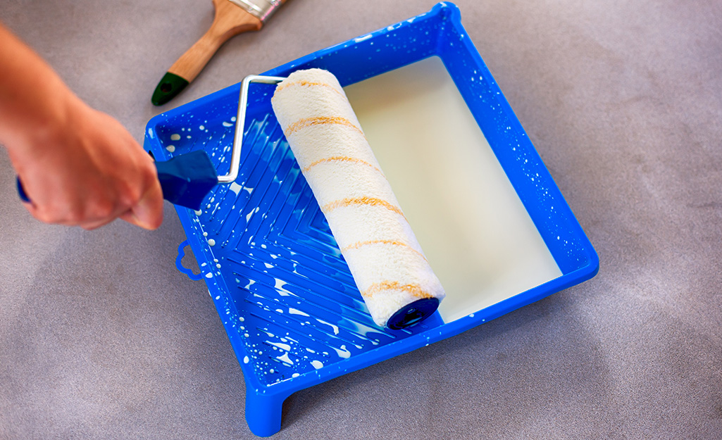 Someone prepares to dip a roller into a tray of cream-colored paint. 