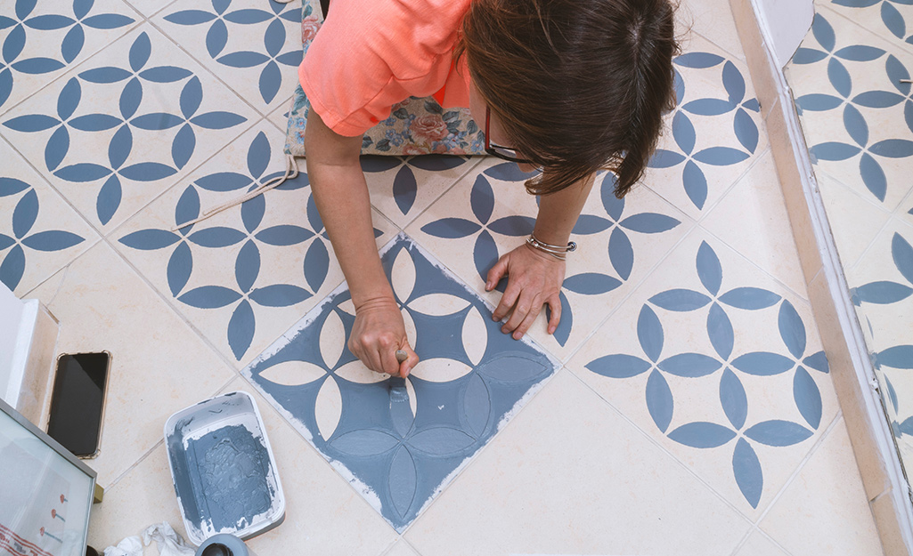 A person uses a stencil to paint a blue pattern on a white tile floor.