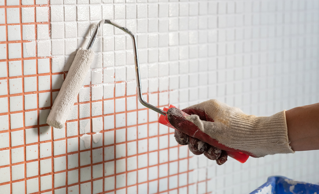A person uses a roller to cover a wall of small tile with paint.