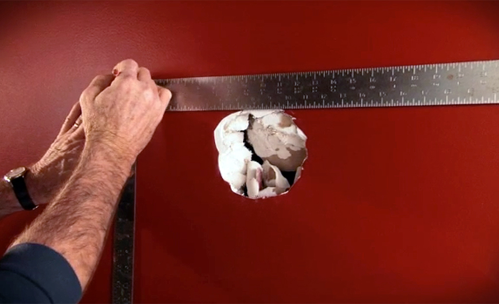 A person using a carpenter's square to measure a large hole in the wall.