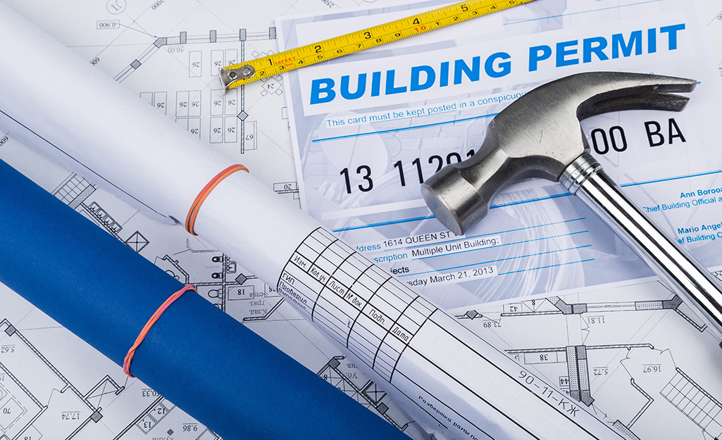 Rolled up building plans, a hammer and a measuring tape lay on top of building permit and architectural drawings.