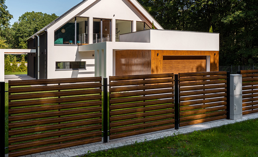 A wood fence with panels made with horizontal boards stands in front of a modern house.