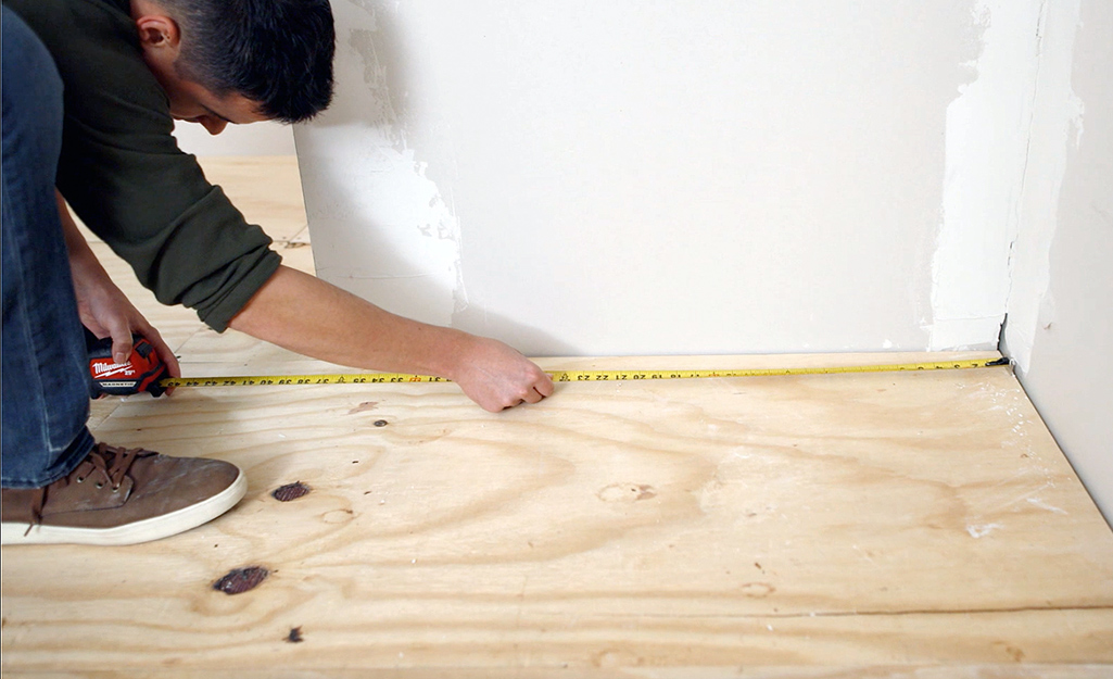 A man kneels as he measures part of a wood subfloor with a yellow measuring tape.