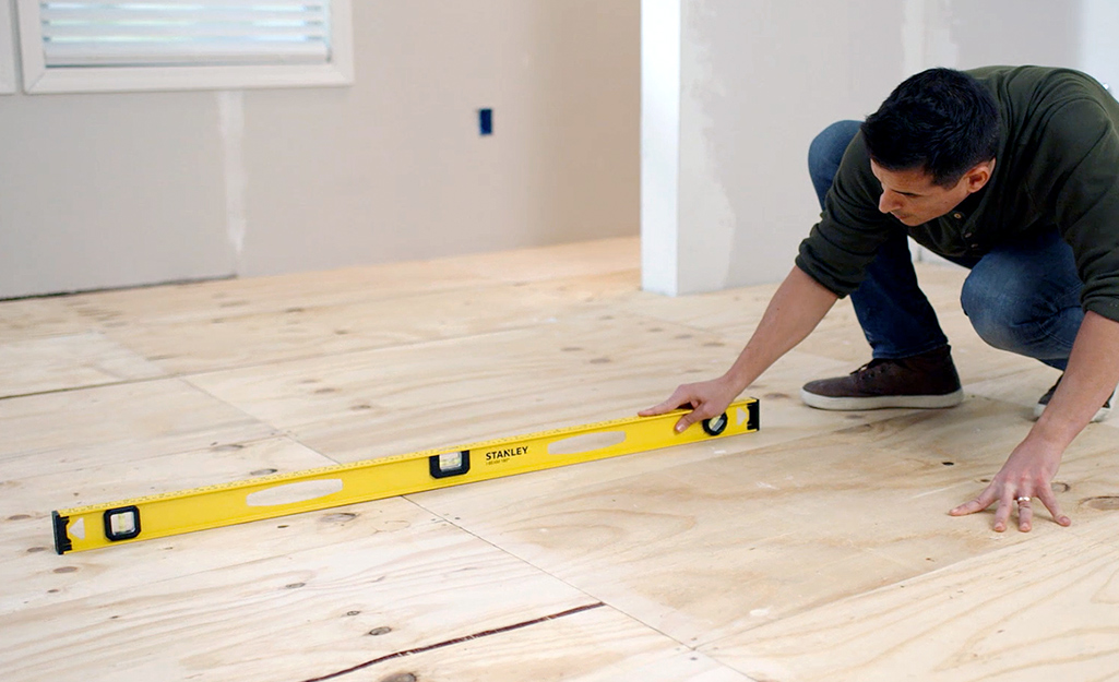 A man uses a yellow level to make sure a wood subfloor is level before tile installation.