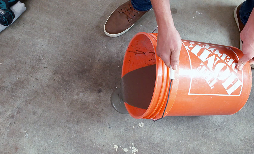 A man pours concrete leveler from an orange Home Depot bucket onto a concrete subfloor.