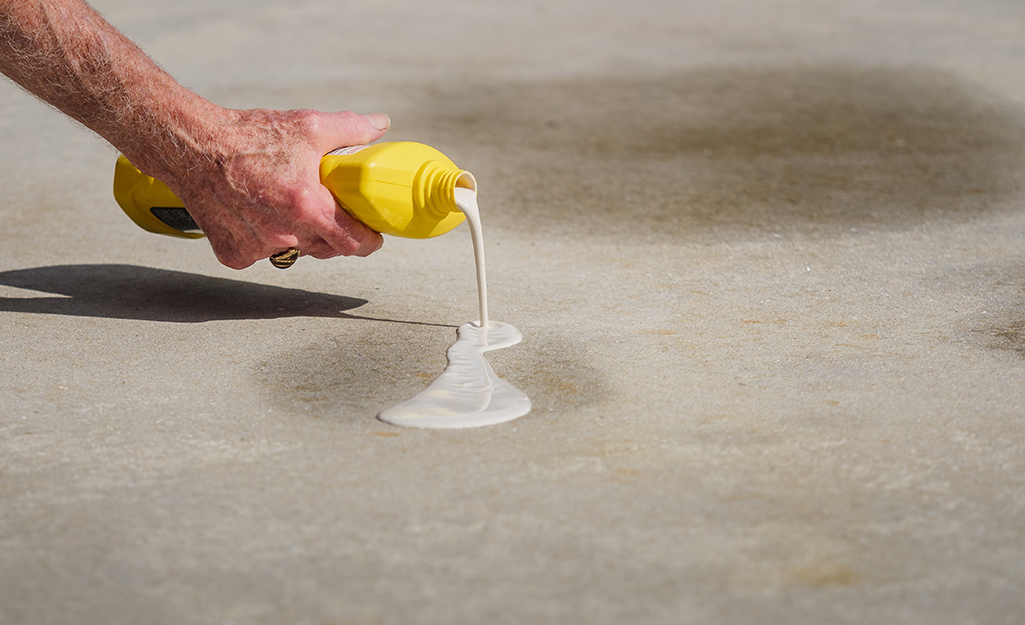 A person pours degreaser from a yellow bottle onto a concrete driveway stain.